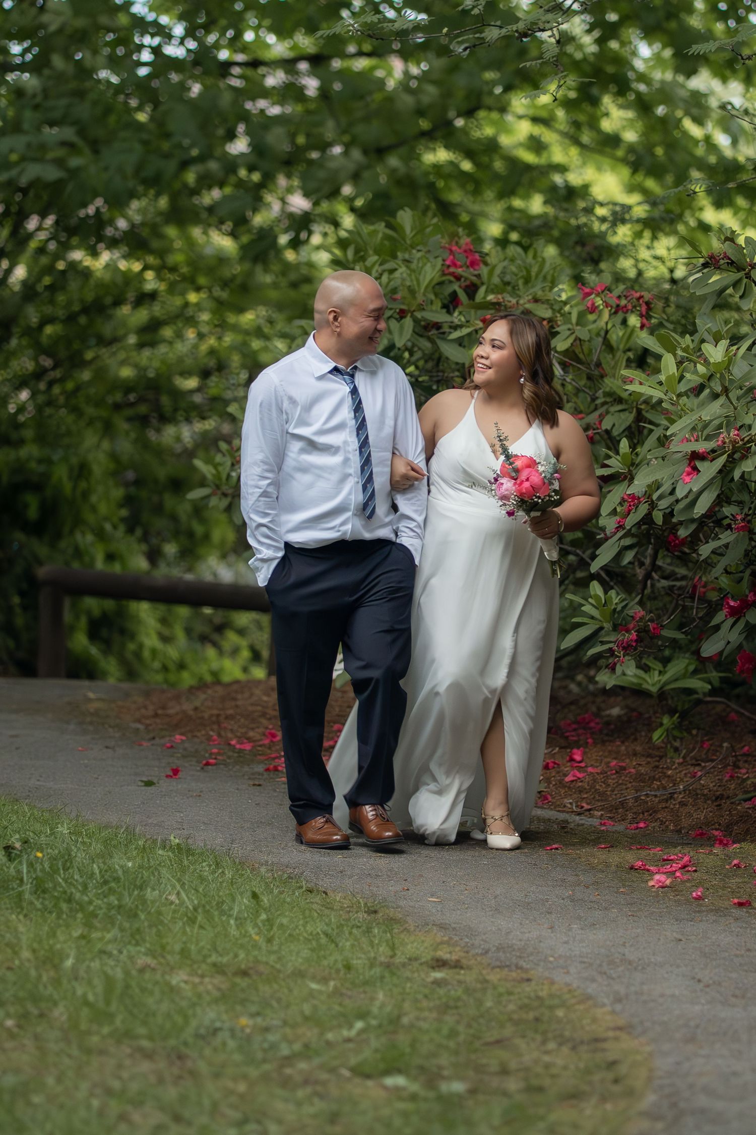 Bride and groom walking on garden path, bride in white gown holding pink bouquet, groom in blue tie and dress shirt.