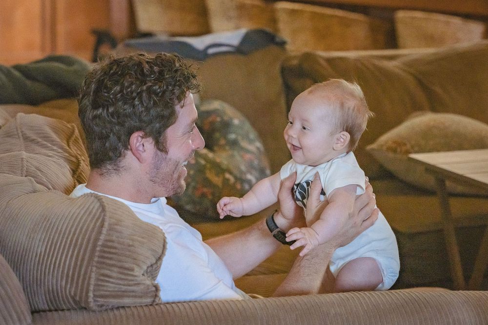 A man in a white shirt smiles at a happy baby sitting on a tan couch in a cozy living room.