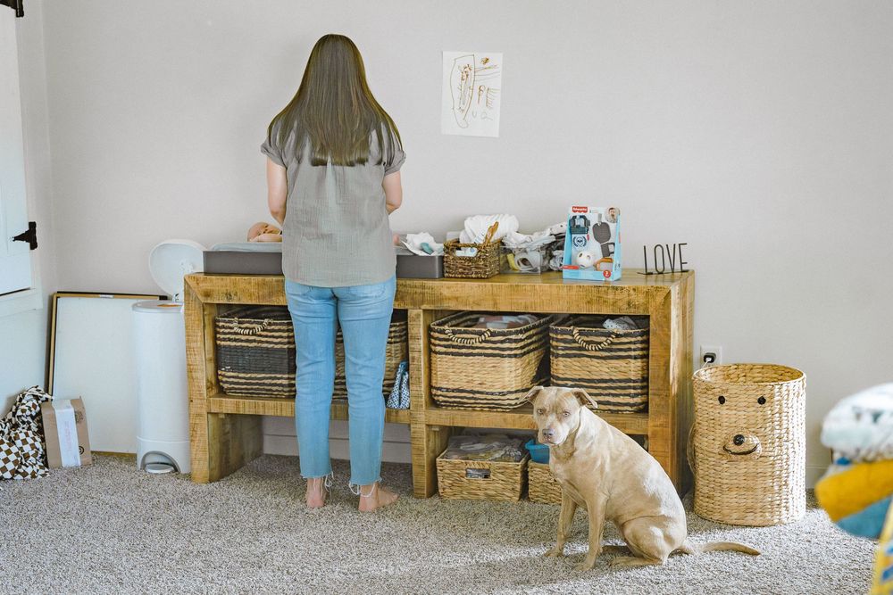Woman standing at rustic wooden console table with wicker baskets, golden retriever sitting on carpet nearby.