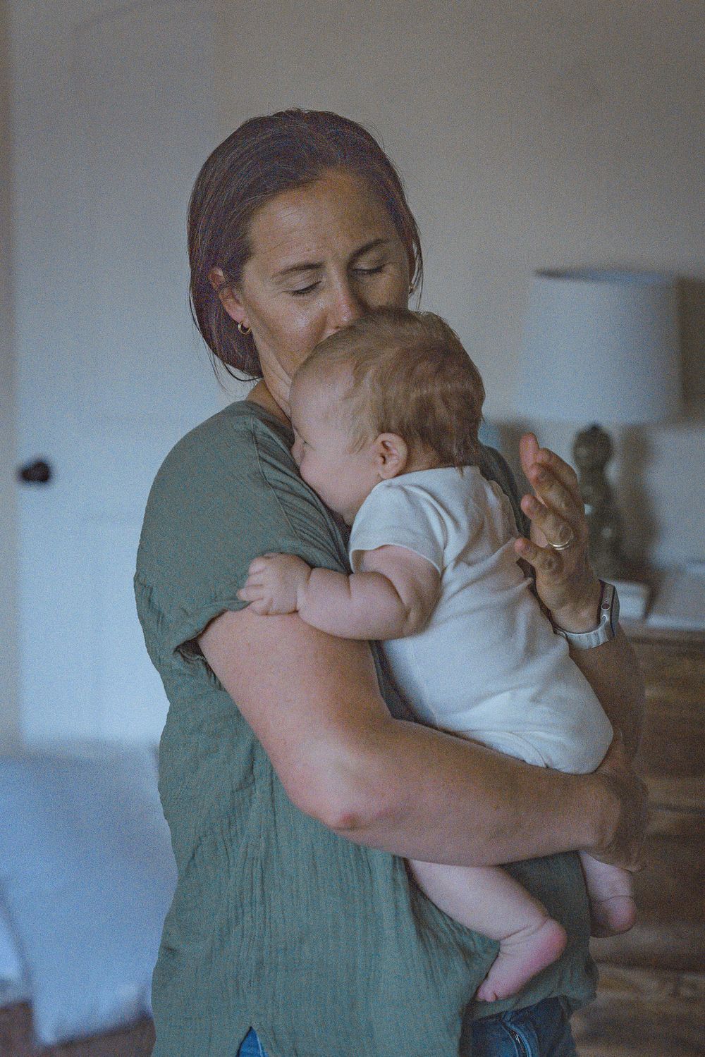 A mother in a green dress tenderly holds and kisses her newborn baby indoors.