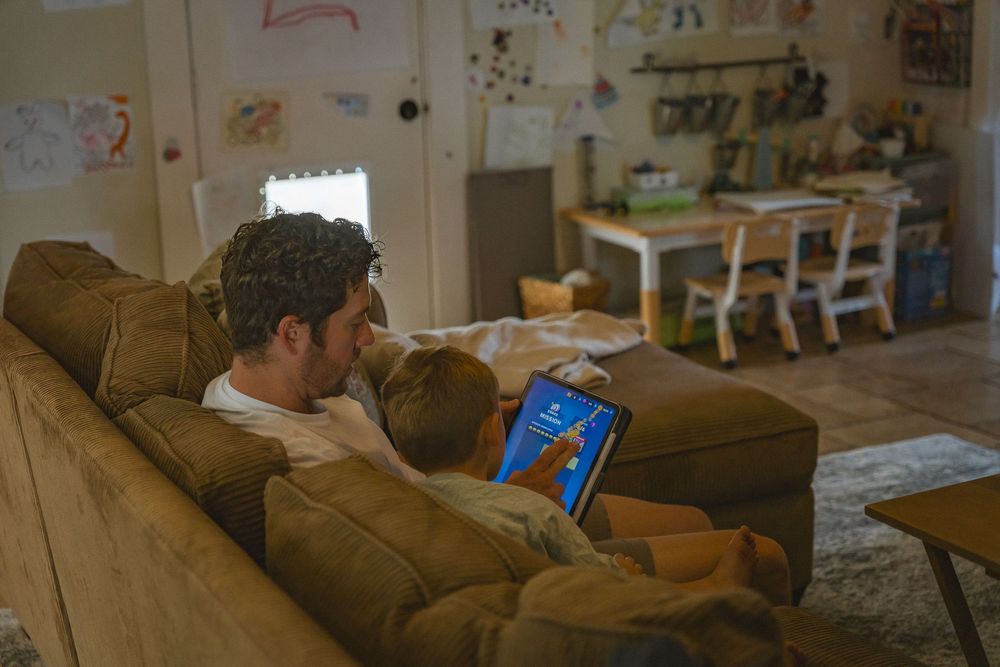 Father and young child sit together on brown sofa watching educational content on a tablet in cozy living room.