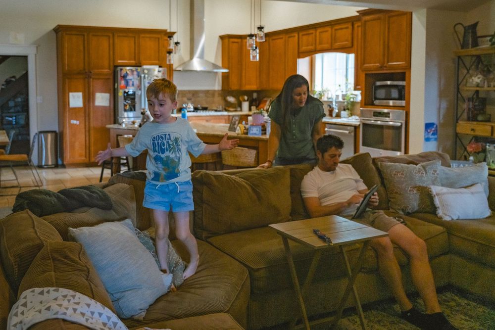 Child jumping on large sectional sofa while two adults relax in open-plan living room with kitchen in background.