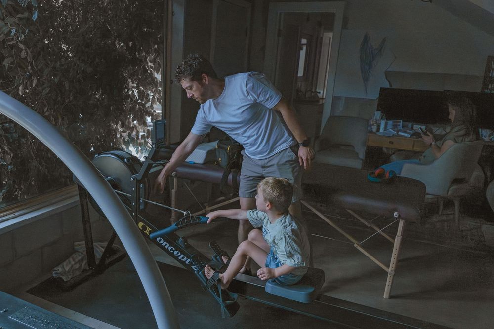 Man and child exercising on rowing machine indoors while woman relaxes on couch in background.