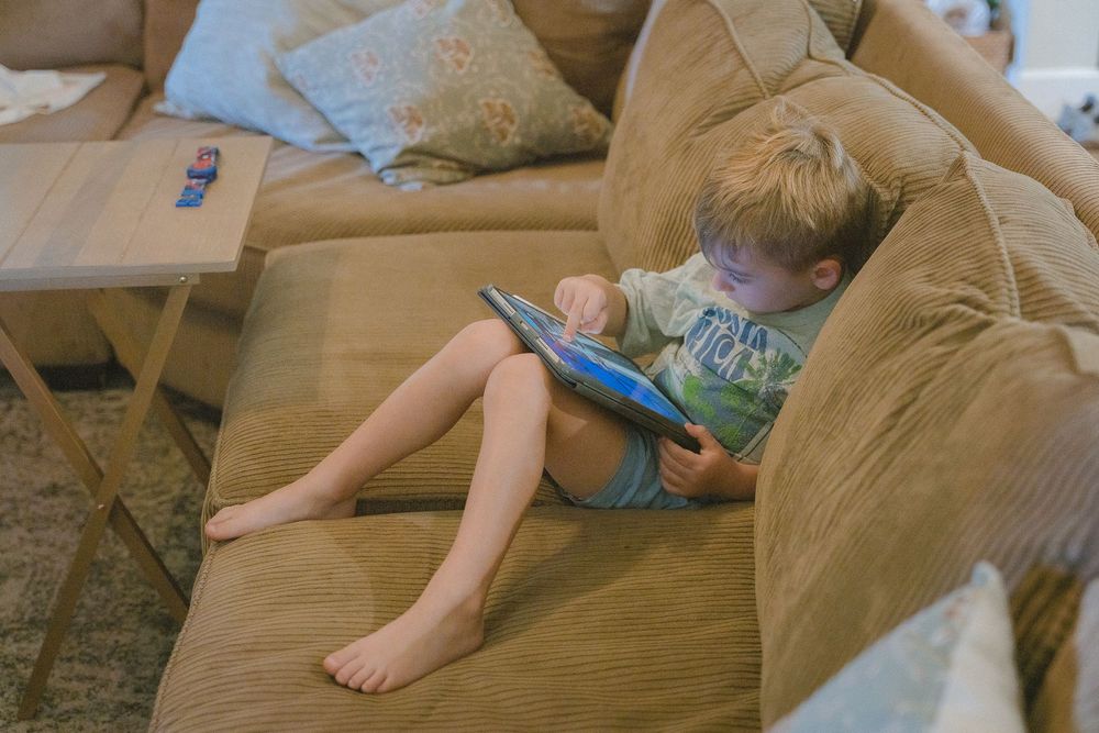 Young barefoot boy sits alone on tan sofa using a tablet computer in a bright living room.