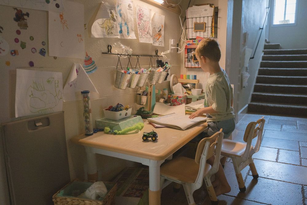 A child sits at a small wooden desk in a cozy room, drawing or writing under a lamp surrounded by art supplies.