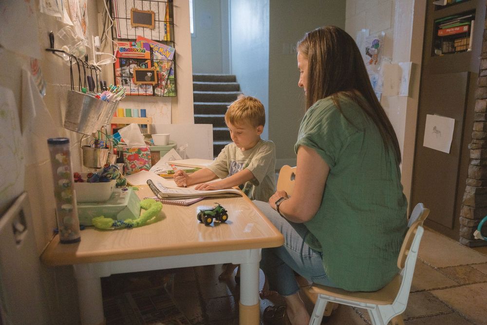 Mother helps young redheaded child with homework at small desk in a cluttered home workspace.