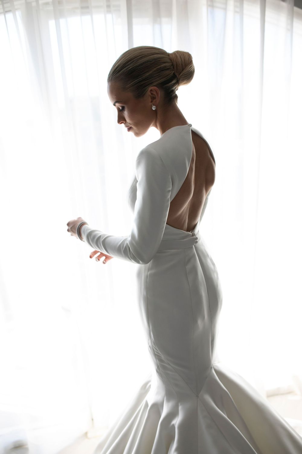 Bride in elegant white backless mermaid wedding gown with long sleeves, hair in updo, standing by sheer curtained window.