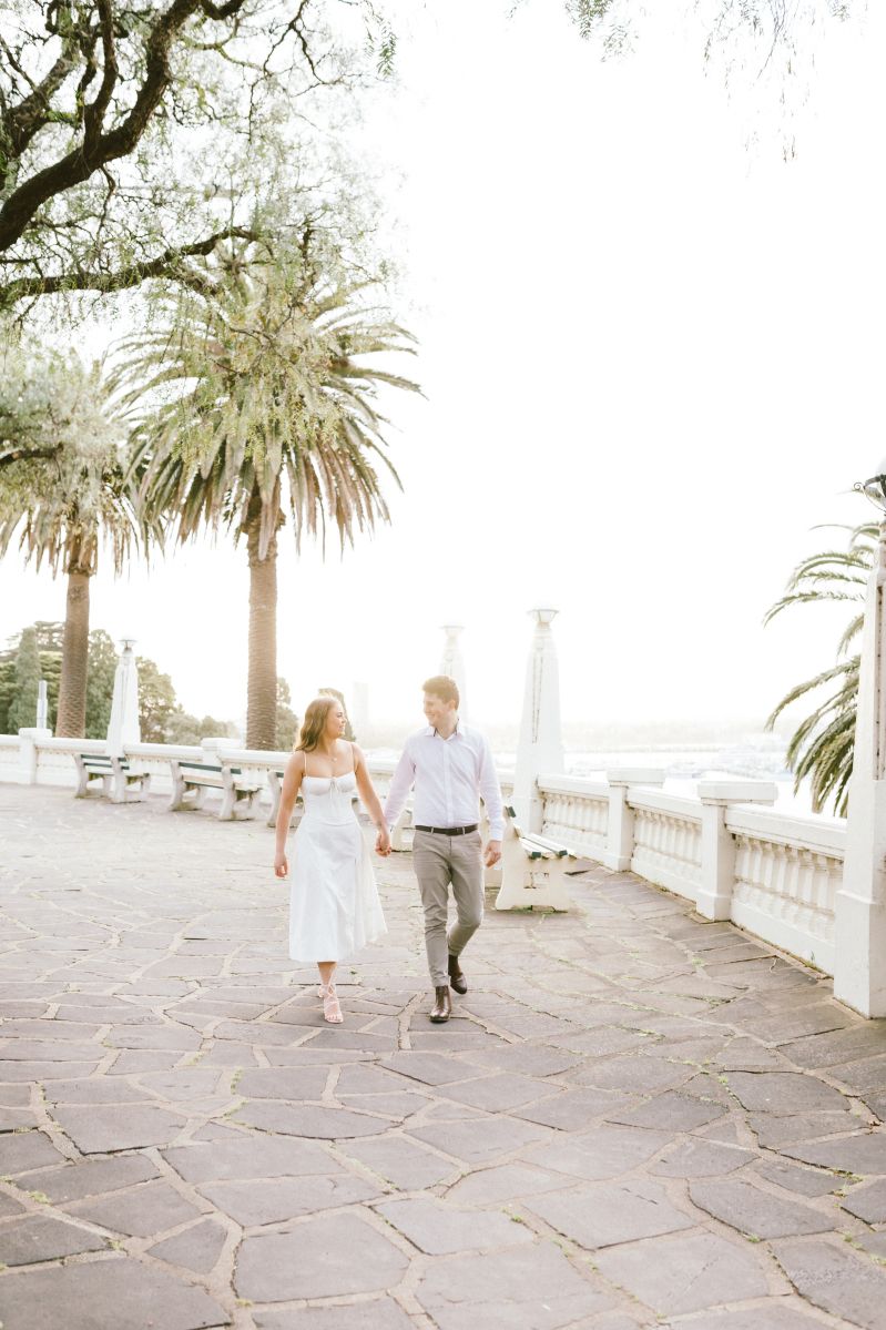 Couple holding hands walking on a stone terrace with palm trees and white balustrade in soft sunlight.