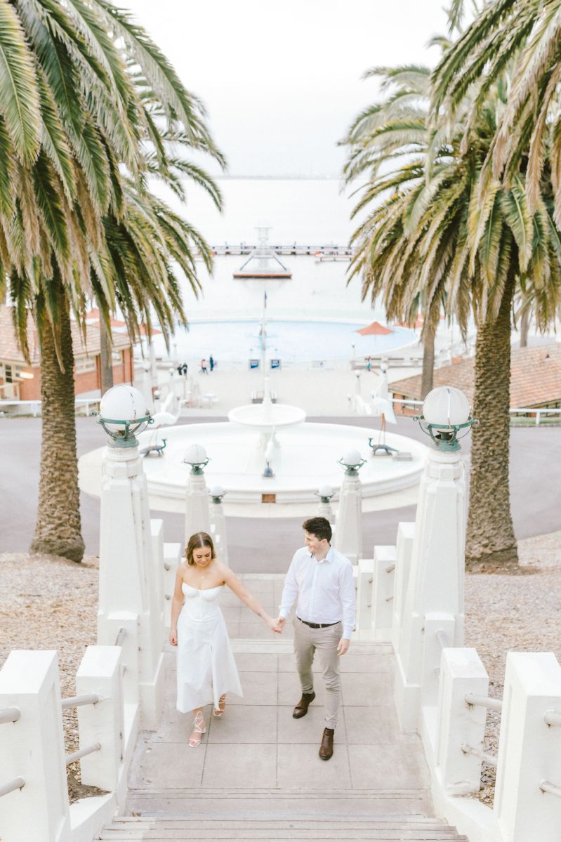 Couple holding hands walking down white stairs flanked by palm trees near a seaside pier.