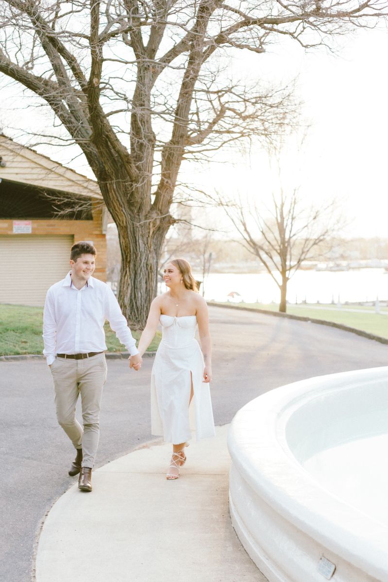 Couple holding hands walking along a waterfront path, woman in white dress and man in gray pants, golden hour light.