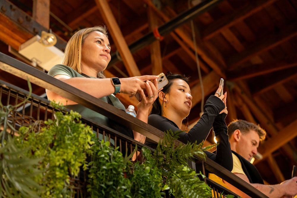 Three people on a balcony using smartphones, surrounded by lush greenery in a rustic wooden venue.