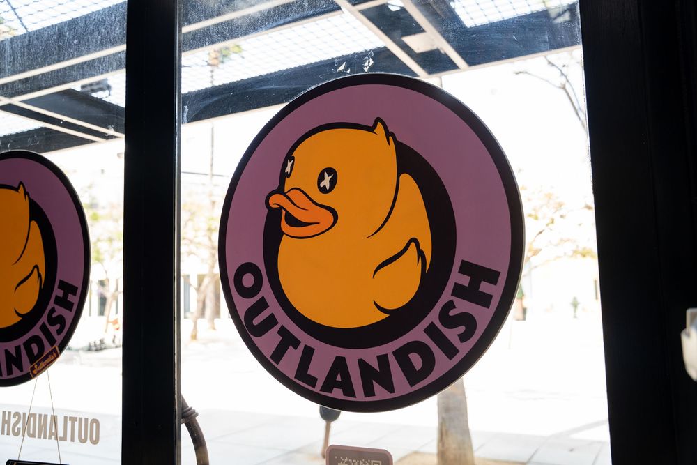Outlandish restaurant logo featuring an orange rubber duck on a circular purple sign displayed on a window.