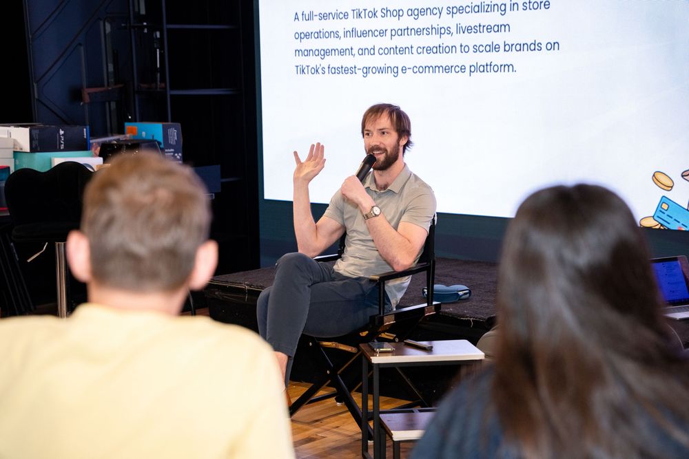 A man speaks to an audience at a TikTok Shop agency presentation, gesturing while seated in front of a projected slide.