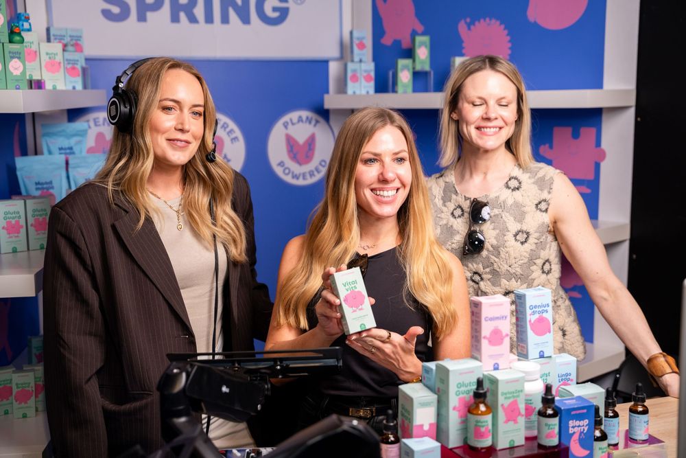 Three women smiling at a spring product display booth with beauty and wellness items on a blue backdrop.