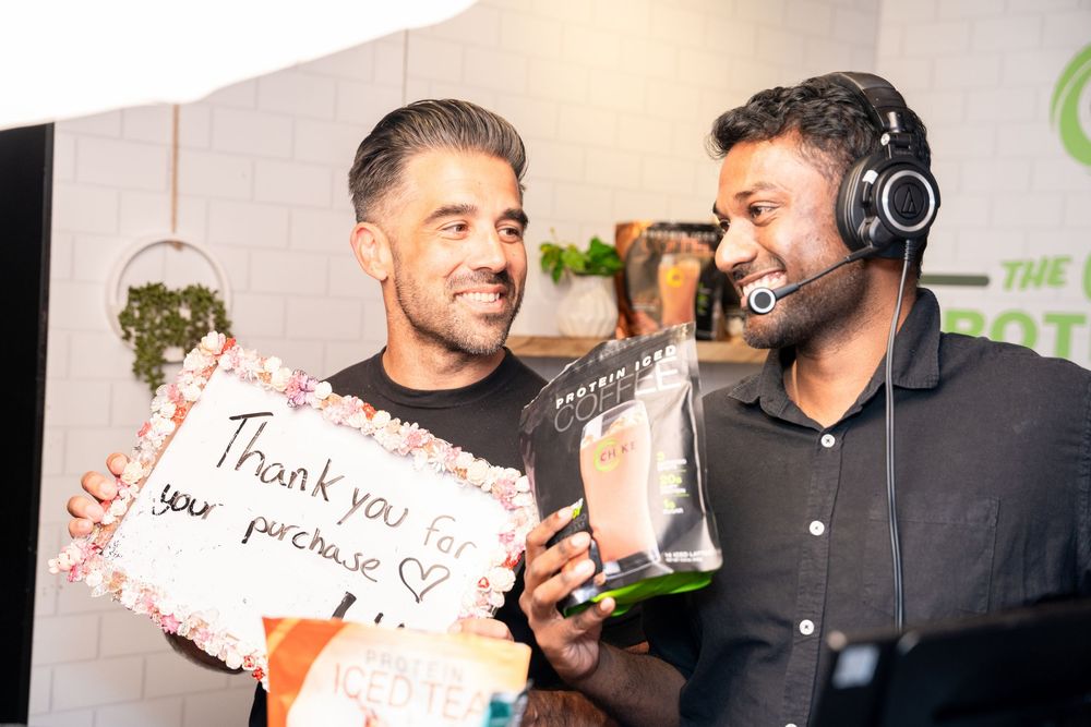 Two men smiling, one holding a thank you for your purchase sign, the other wearing a headset microphone indoors.