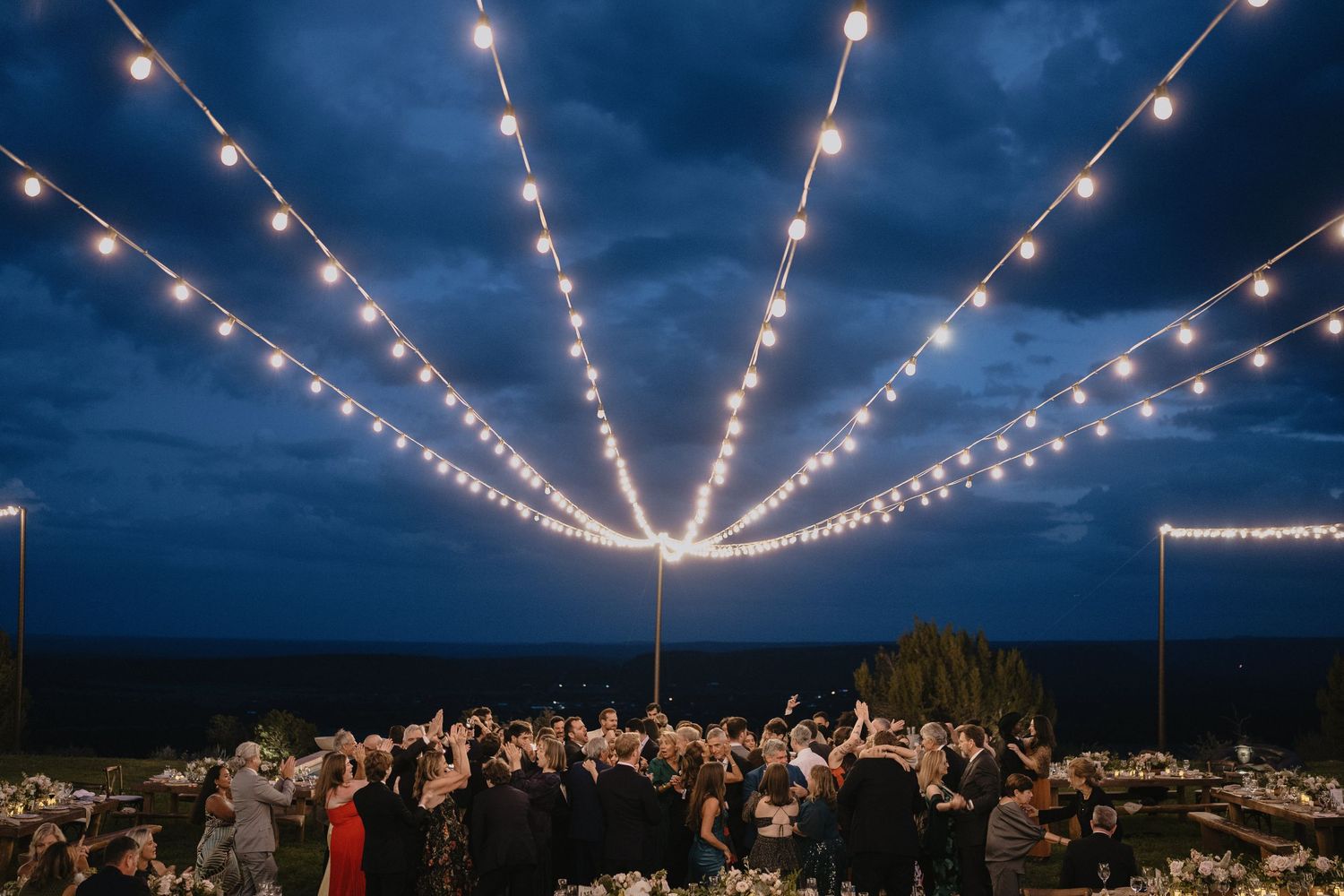 Outdoor wedding reception at dusk with string lights radiating overhead, guests gathered under glowing bulbs against a dramatic blue sky.