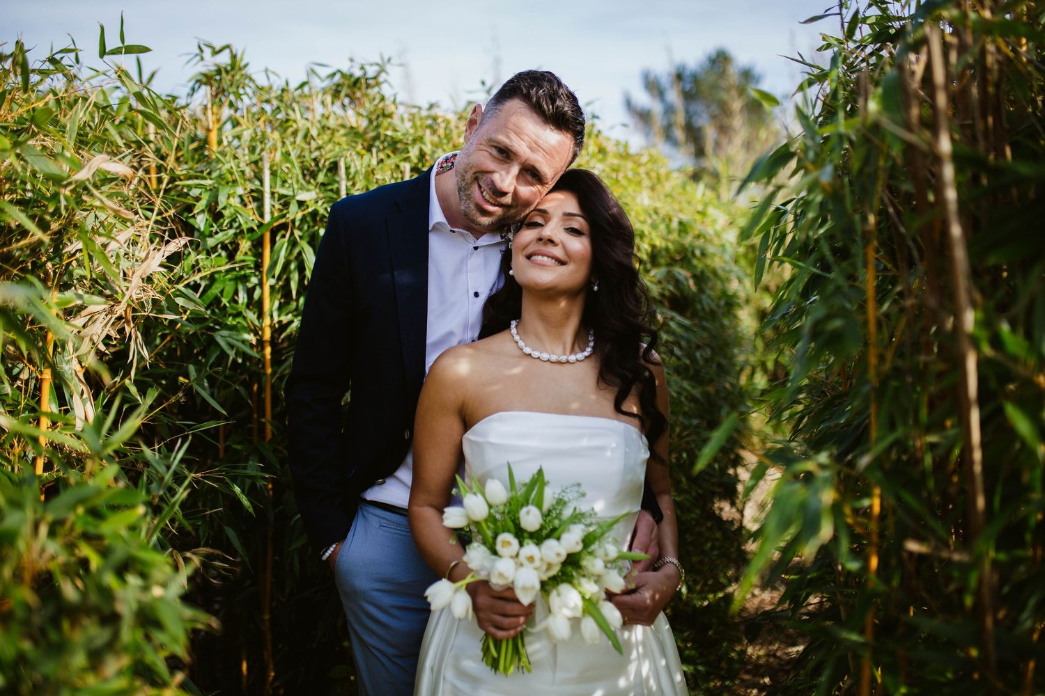 Bride and groom share a romantic moment surrounded by lush green bamboo, bride holding white tulip bouquet.