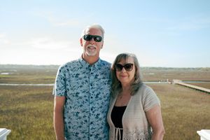 Smiling couple posing outdoors near a coastal marsh with a wooden boardwalk and scenic wetlands in the background.