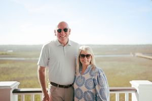 Smiling couple posing on a sunny deck overlooking a coastal marsh landscape.