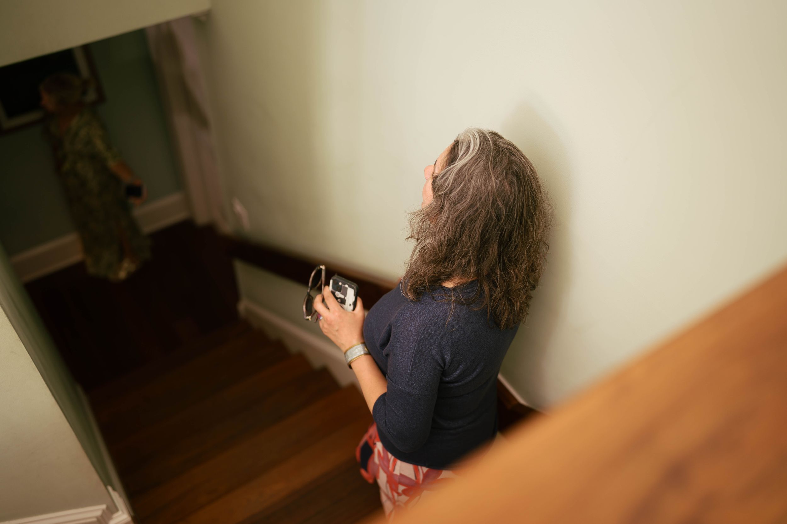 Woman with gray hair holds camera, photographing someone on staircase landing in a warmly lit home.