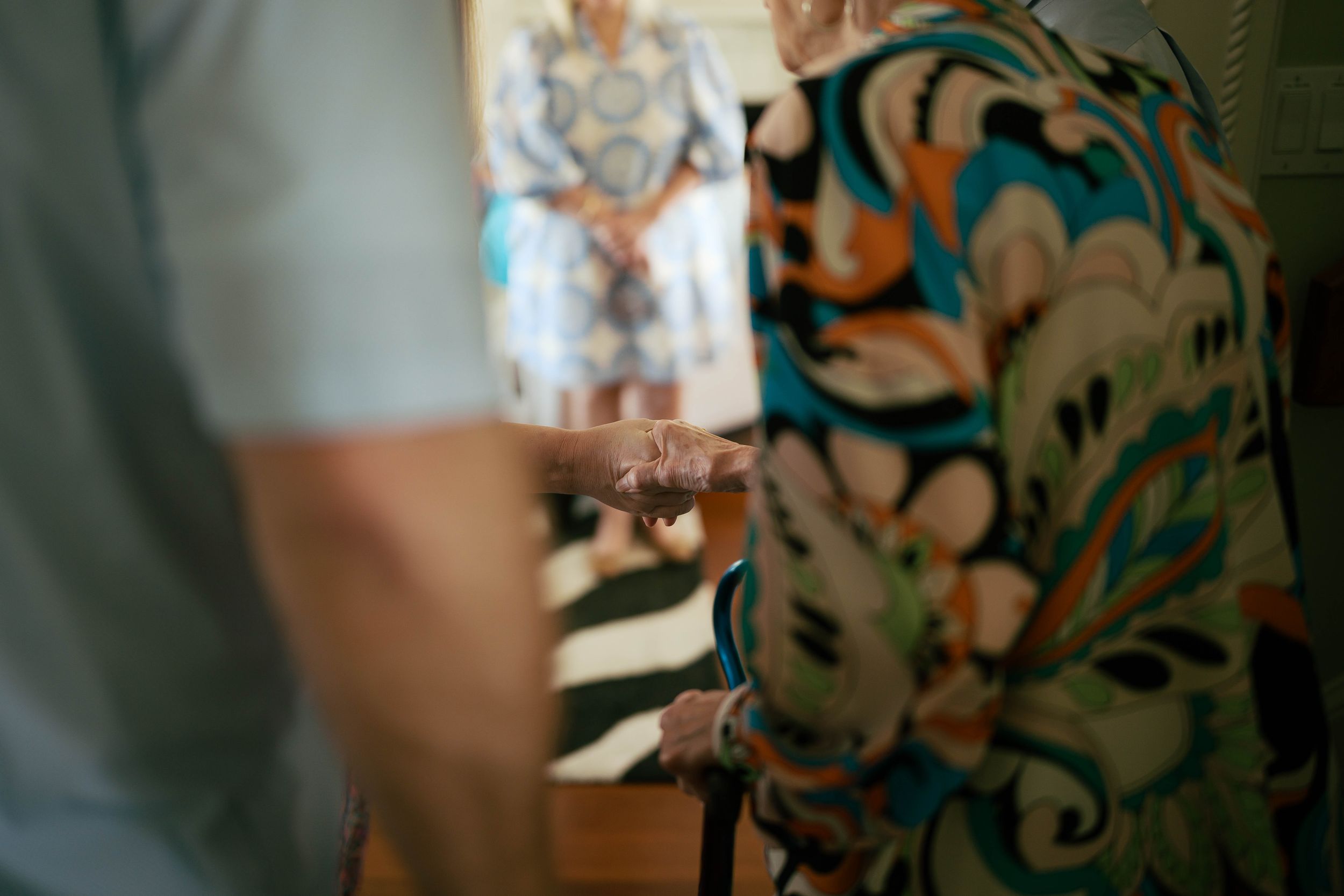 Elderly woman in colorful floral dress holding a cane at an indoor gathering with guests in background.