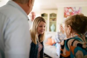 Young blonde woman smiling and conversing with elderly guests at an indoor family gathering.