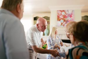 Elderly adults socializing indoors, with a bald man shaking hands and exchanging gifts in a bright living room.