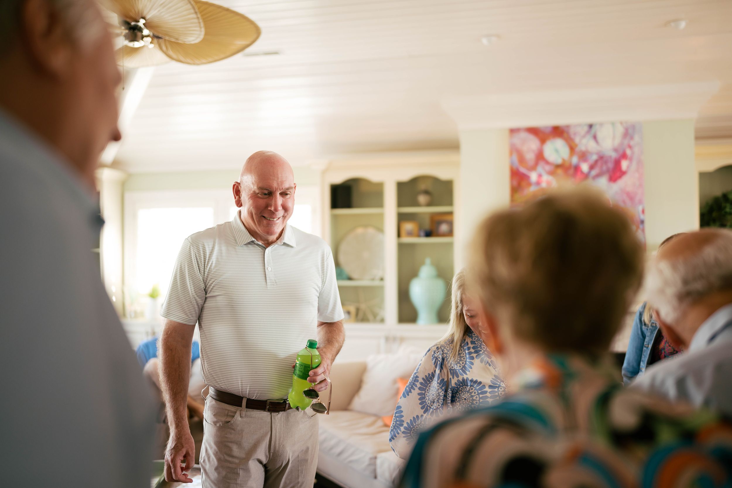 Smiling senior man holding a drink socializes with elderly friends in a bright, cozy living room.