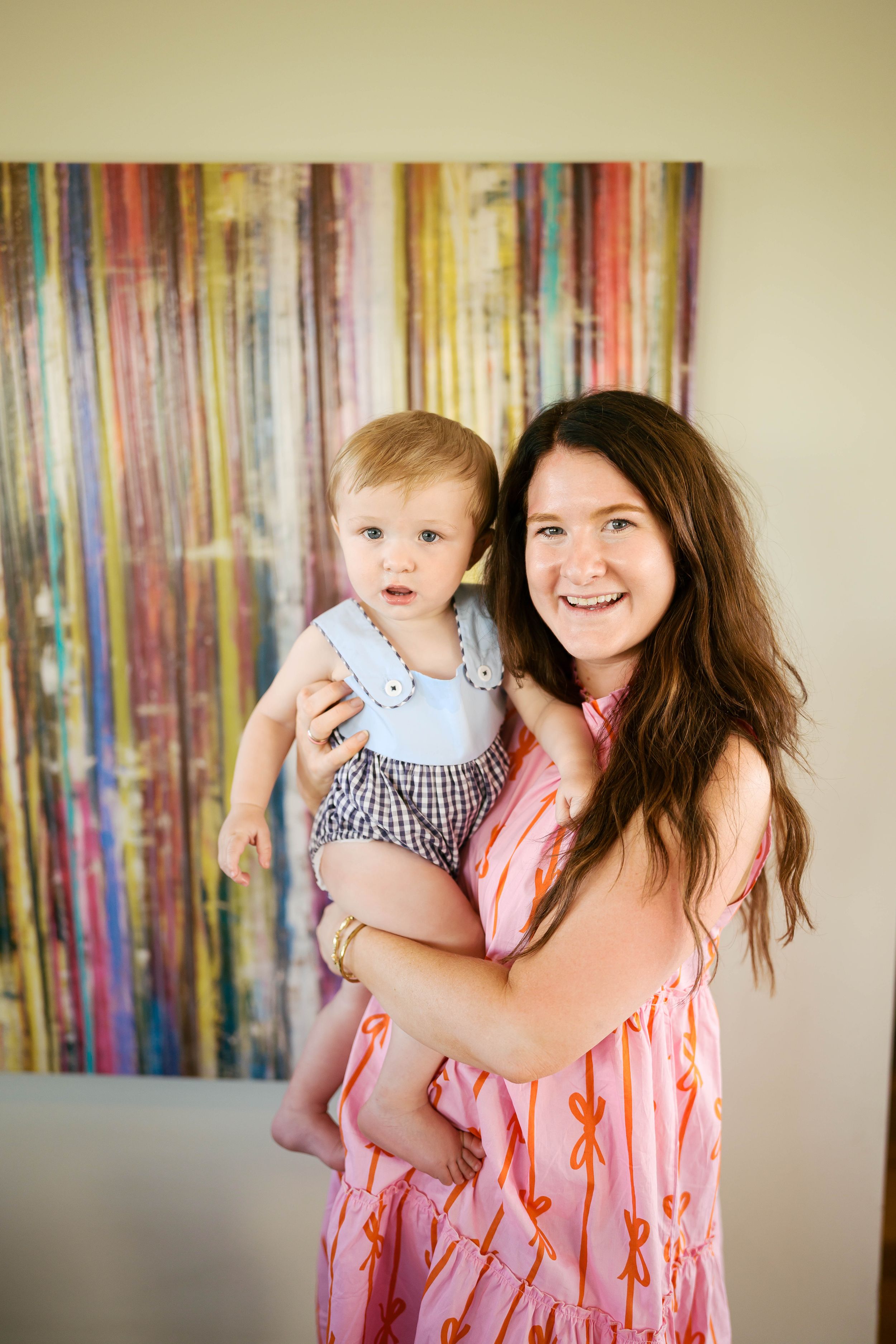Smiling young woman in pink dress holding a baby in blue outfit in front of colorful abstract painting.