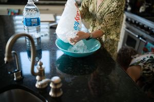 Woman pouring ice from a bag into a teal bowl on a dark granite kitchen counter near a sink.