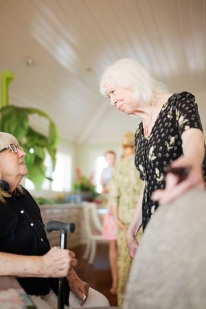 Two elderly women conversing indoors, one seated with a cane, the other standing in a floral dress, with people in the background.