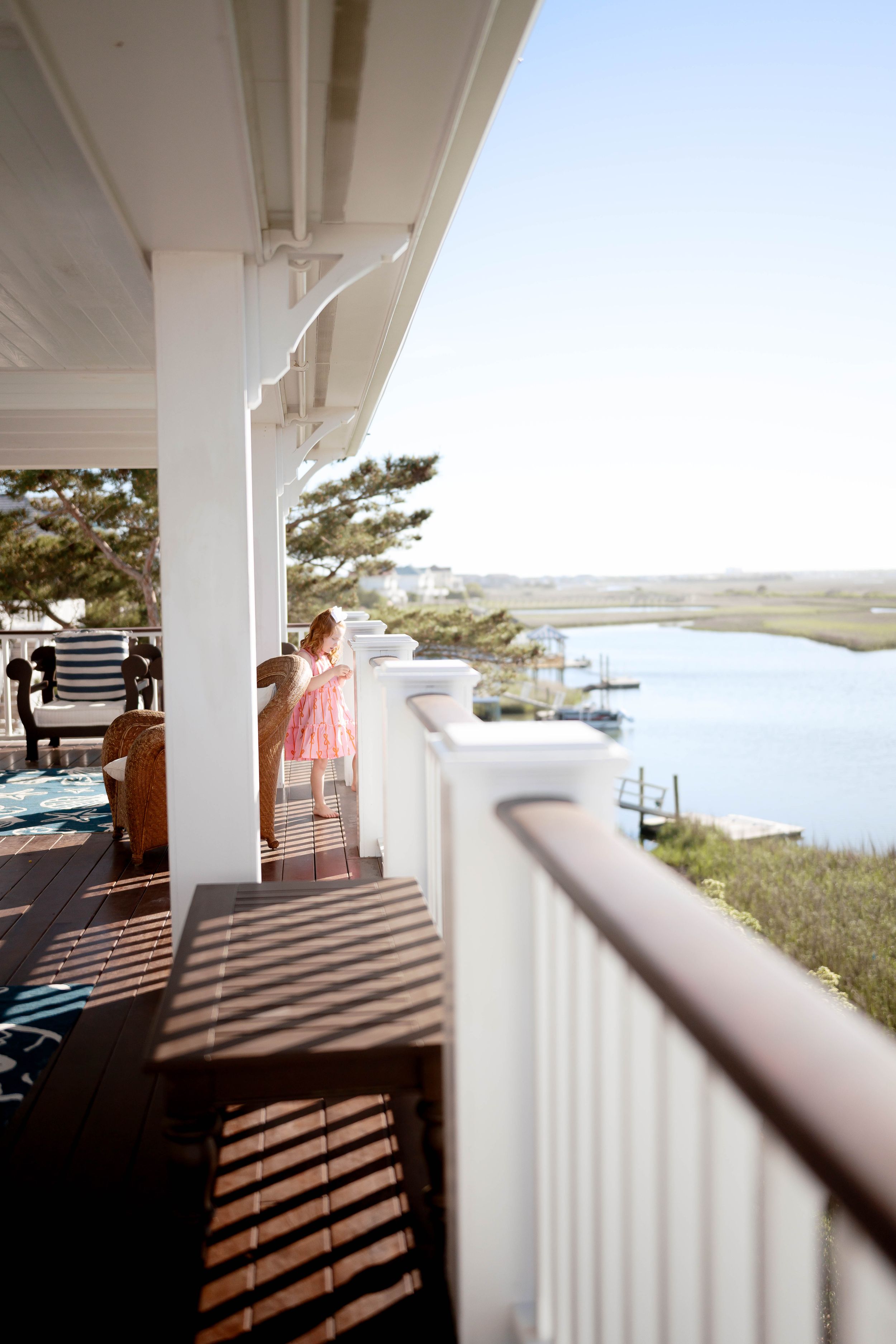 A girl in a pink dress stands on a white porch railing overlooking a serene coastal waterway on a sunny day.