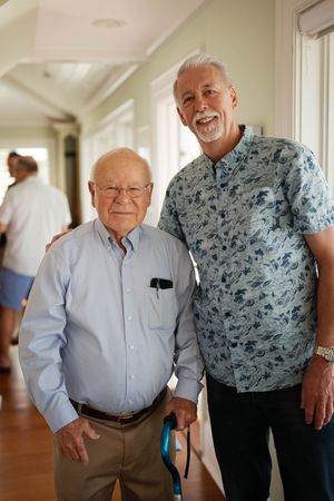 Two elderly men smiling together indoors, one holding a cane, wearing casual button-up shirts in a bright hallway.