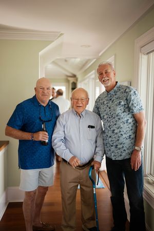 Three men smiling together indoors, elderly man with cane flanked by two middle-aged men in blue shirts.