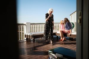 Three women socializing on a sunny rooftop deck, one standing in a floral dress while two others sit nearby.