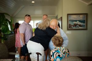 Elderly man and woman share a warm hug indoors surrounded by family members in a bright coastal home.
