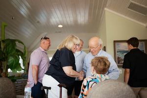 Group of elderly adults gathered indoors, sharing a warm conversation in a bright, tropical-style room.