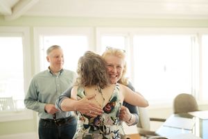 Two women share a warm hug indoors while a smiling man stands in the background near bright windows.