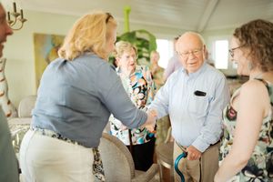 Elderly man shaking hands with a blonde woman at a social gathering, with other guests standing nearby in a bright room.