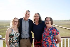 Four smiling adults posing together outdoors on a sunny day with a scenic marsh landscape in the background.