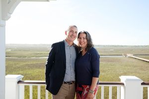 Smiling couple posing together on a white porch overlooking a scenic coastal marsh landscape.