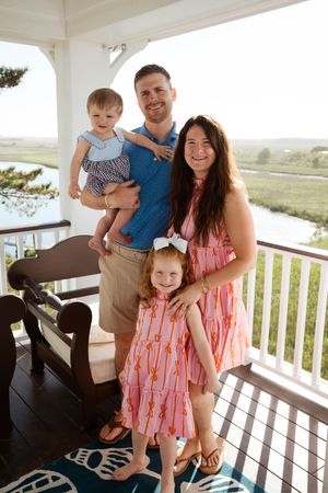 Happy family of four posing on a porch overlooking a marsh, wearing colorful summer outfits on a sunny day.