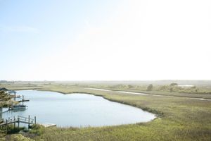 Aerial view of a serene coastal marsh with a small pond, wooden dock, and expansive wetlands under a bright sky.