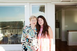 Two women smiling together on a sunny porch, one older in a floral blouse and one younger in a pink striped top.
