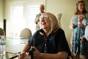 Elderly woman with glasses laughing joyfully while seated, with guests standing in bright room background.