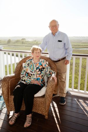 Elderly couple posing on a sunny deck, woman seated in wicker chair, man standing beside her with marsh views.