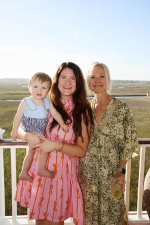 Two women and a baby girl pose on a balcony overlooking a marsh, wearing colorful summer dresses.