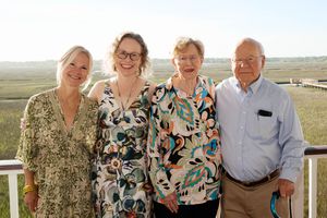 Four people smiling together on an outdoor deck overlooking a scenic marsh landscape on a sunny day.