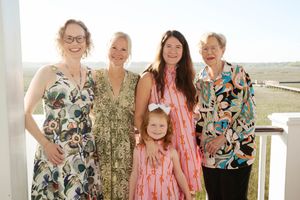 Five women and a young girl in colorful summer dresses pose together smiling on a sunny outdoor balcony.