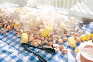 A classic seafood boil with shrimp, corn, and potatoes spread on a blue checkered tablecloth outdoors.